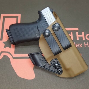 May include: A black handgun in a tan holster with black clips. The holster is on a dark surface with a Texas outline logo. The text "Texas Holsters" is visible in the background.