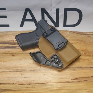May include: A tan leather holster with a black plastic clip and a black handgun inside. The holster is designed to be worn on a belt.