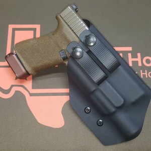 May include: A black leather holster with a brown and black handgun inside. The holster has two adjustable straps and is secured with screws. The holster is on a brown background with the text "Holsters" printed in pink.