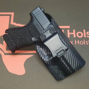 May include: A black handgun in a black carbon fiber holster with a black clip. The holster has two black screws on the bottom and the clip has the text "1.5" Made in U.S.A."