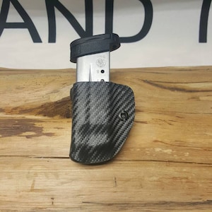 May include: A black carbon fiber magazine holder with a silver magazine inside. The magazine has the text "9mm" and a logo on it.