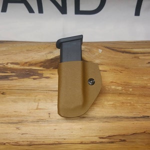 May include: A tan leather magazine holster with a black magazine inside. The holster is on a wooden surface.