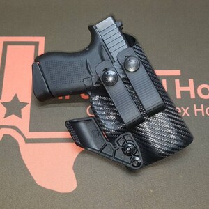 May include: A black handgun in a black carbon fiber holster with a black belt clip. The holster has a black adjustable retention screw. The holster is on a green background with a red outline of the state of Texas.