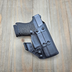 May include: A black handgun with a textured grip is holstered in a black leather holster with a clip. The holster has two metal clips on the back.