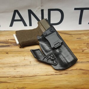 May include: A black and grey carbon fiber holster with a black clip and a brown and silver handgun inside.