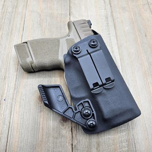 May include: A black holster with a black clip and a tan handgun. The holster is made of a durable material and features a secure design. The handgun is partially visible, showing its grip and slide.