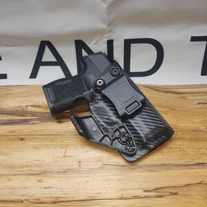 May include: A black handgun in a black carbon fiber holster. The holster has a belt clip and a release button.