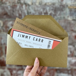 K&ouml;nnte beinhalten: Ein brauner Umschlag mit einem Ticket f&uuml;r eine Jimmy Carr-Show. Das Ticket ist wei&szlig; und korallenfarben mit schwarzem Text, einschlie&szlig;lich Veranstaltungsort und Datum. Der Umschlag wird vor einer Backsteinmauer gehalten.