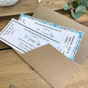 Könnte beinhalten: Zwei Geschenkgutscheine in einem braunen Umschlag. Die Gutscheine haben goldene Akzente und den Text "You're on the Nice List" und "A trip to The Garage Spa". Weihnachtsdekorationen und Grünzeug sind im Hintergrund.