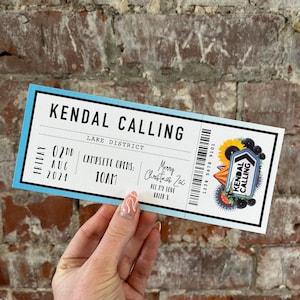 K&ouml;nnte beinhalten: Ein rechteckiges Ticket mit blauem und wei&szlig;em Design, das den Text "KENDAL CALLING" und Veranstaltungsdetails enth&auml;lt. Das Ticket enth&auml;lt einen Barcode, ein Veranstaltungslogo und handschriftliche Notizen. Gehalten vor einer Backsteinmauer.