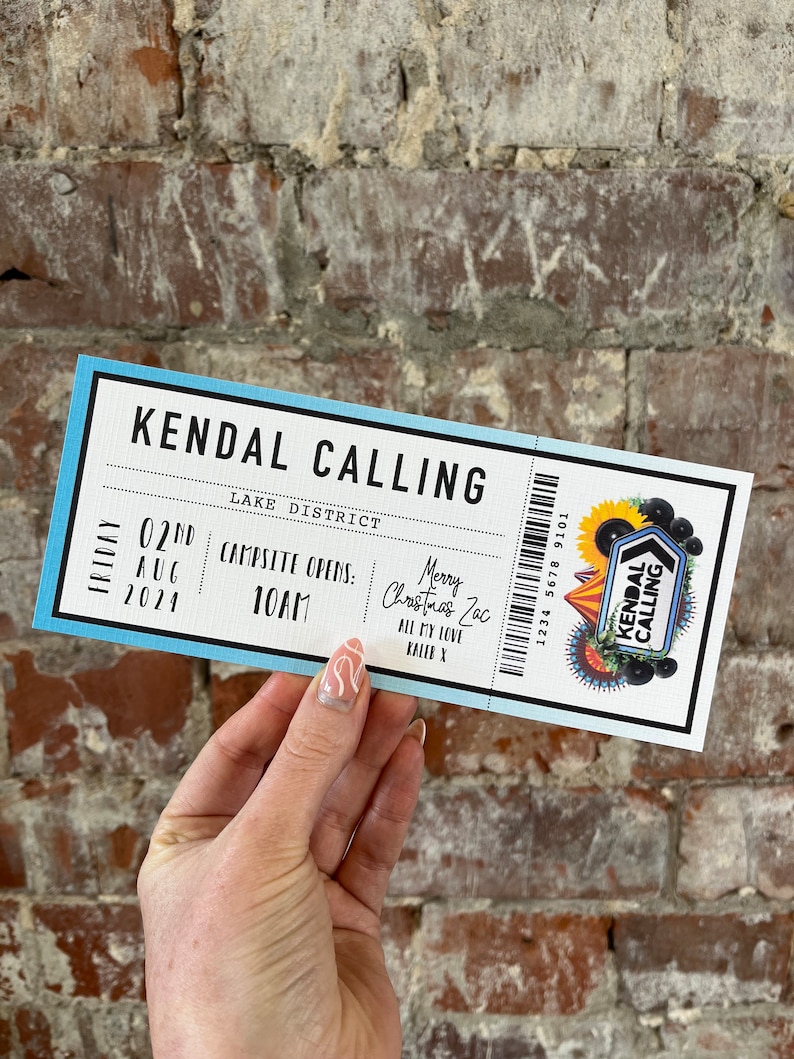 K&ouml;nnte beinhalten: Ein rechteckiges Ticket mit blauem und wei&szlig;em Design, das den Text "KENDAL CALLING" und Veranstaltungsdetails enth&auml;lt. Das Ticket enth&auml;lt einen Barcode, ein Veranstaltungslogo und handschriftliche Notizen. Gehalten vor einer Backsteinmauer.