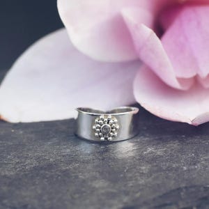 Puede incluir: Un anillo plateado con un diseño floral, sobre una piedra gris oscuro y una flor rosa suave. El anillo presenta un grupo de pequeñas piedras oscuras en el centro, creando un aspecto delicado y elegante.