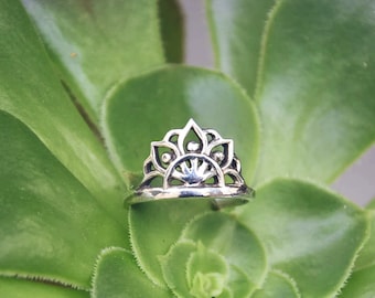 Könnte beinhalten: Ein silberner Ring mit einem filigranen, kunstvollen Design, das an eine Krone oder einen Sonnenstrahl erinnert. Der Ring liegt auf einer grünen Sukkulente.