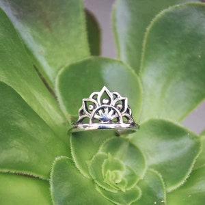 Peut inclure: Une bague en argent avec un design délicat et complexe ressemblant à une couronne ou un soleil éclatant. La bague est posée sur une plante succulente verte.