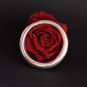 Peut inclure: Un bracelet en argent avec une surface lisse et arrondie. Le bracelet repose sur une rose rouge.