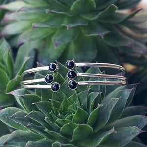Peut inclure: Un bracelet jonc argenté composé de trois bandes et de six pierres précieuses noires. Le bracelet est photographié sur une plante succulente verte.