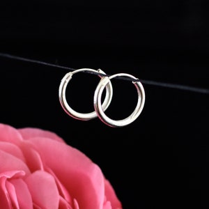 Peut inclure: Une paire de boucles d'oreilles créoles en argent suspendues à une ficelle noire sur un fond noir. Une rose rose est au premier plan.