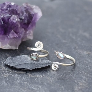 Könnte beinhalten: Zwei verstellbare, silberfarbene Ringe mit tropfenförmigen Edelsteinen und Spiralakzenten. Ein großer, violetter Amethystkristall liegt hinter den Ringen auf einer dunkelgrauen Oberfläche. Die Ringe sind wahrscheinlich für die Finger oder Zehen.