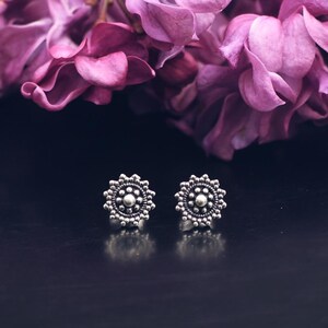 Peut inclure: Une paire de boucles d'oreilles en argent avec un motif floral, sur fond sombre. Les boucles d'oreilles sont au point, avec un bouquet de fleurs violettes en arrière-plan. Les boucles d'oreilles ont un motif circulaire détaillé.