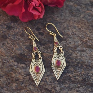 Peut inclure: Une paire de boucles d'oreilles pendantes dorées avec un motif géométrique et de petites pierres précieuses rouges ovales. Les boucles d'oreilles sont présentées sur une surface grise texturée, avec une rose rouge en arrière-plan.