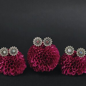 Peut inclure: Trois paires de boucles d'oreilles en argent avec des pierres de couleurs différentes serties dans un motif circulaire. Les boucles d'oreilles sont présentées sur trois petits supports en forme de fleur rouge.