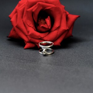 May include: A silver ring with a distinctive double-band design, with a small, round detail. A vivid red rose is behind the ring, set against a dark grey background. The image highlights the ring's modern style and the contrast with the flower.