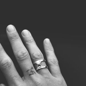 May include: A silver ring with a distinctive design, incorporating a small circular feature at its centre. The ring is worn on a finger, set against a dark backdrop. The ring's style is modern and minimalist.