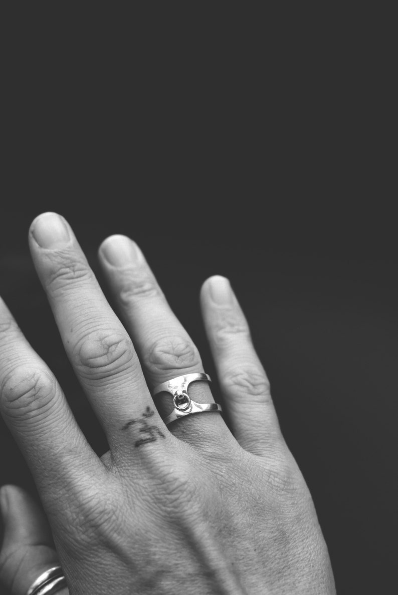 May include: A silver ring with a distinctive design, incorporating a small circular feature at its centre. The ring is worn on a finger, set against a dark backdrop. The ring's style is modern and minimalist.