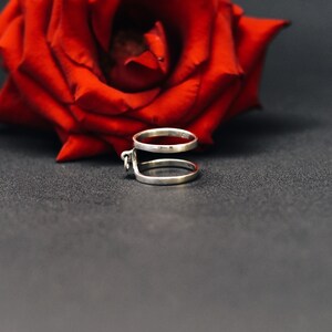 May include: A silver ring featuring two bands joined by a small decorative detail. A vivid red rose is in the background, set against a dark grey surface. The ring's design is modern and minimalist.