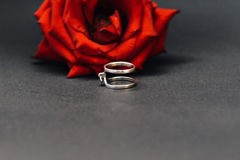 May include: A silver ring featuring two bands joined by a small decorative detail. A vivid red rose is in the background, set against a dark grey surface. The ring's design is modern and minimalist.