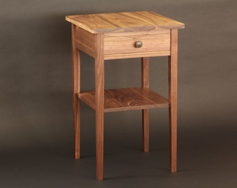 May include: A wooden side table with a drawer and a lower shelf. The table is made of a light brown wood and has a square top. The drawer has a round, brass-colored knob. The table is approximately 28 inches tall.