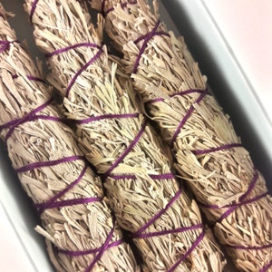 May include: Three bundles of white sage smudge sticks tied with purple string. The sticks are arranged in a row, with the ends of the sticks visible.