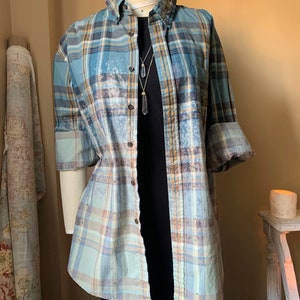 May include: A blue, tan, and white plaid button-down shirt with rolled-up sleeves is displayed on a mannequin. The shirt is worn over a black dress and a crystal pendant necklace. The shirt has small, scattered embellishments.