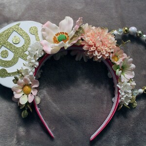 Op de afbeelding: Een paar roze en witte bloemenhoofdbandoren met een witte cirkel met een gouden glitterontwerp. De hoofdband is versierd met witte en roze bloemen, groene bladeren en gouden kralen.