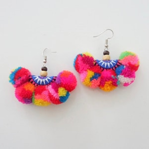 May include: A pair of colorful pom-pom earrings with a blue and white embroidered design. The earrings are made with a variety of colors, including pink, red, yellow, green, and blue.