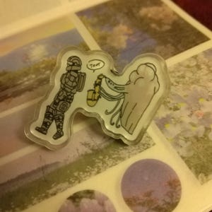 May include: A clear plastic pin with a cartoon illustration of a space-suited figure handing a saxophone to a large, gray, blob-like creature. The creature has a speech bubble that says "Toot".