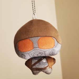 May include: A plush keychain featuring a character with a brown helmet, bright orange visor, and a gray gas mask design. The figure has a small body with brown and gray details, including a strap across the chest, suspended by a silver ball chain.