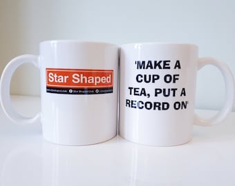 May include: Two white ceramic mugs. One mug has the text "Star Shaped" in a red rectangle with a website address and social media icons. The other mug has the text "'MAKE A CUP OF TEA, PUT A RECORD ON'" in black.