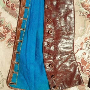 May include: A brown leather corset with a blue lining and button closures. The corset is open and laid flat on a floral patterned fabric.