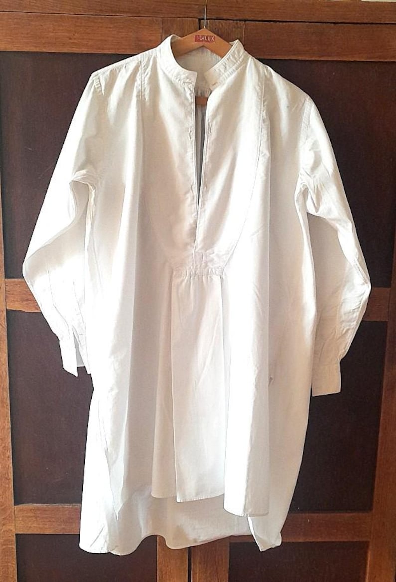 K&ouml;nnte beinhalten: Ein langes, wei&szlig;es Hemd im Tunika-Stil mit Stehkragen und langen &Auml;rmeln. Das Hemd hat eine Knopfleiste und eine lockere, flie&szlig;ende Passform. Das Kleidungsst&uuml;ck h&auml;ngt an einem Holzb&uuml;gel.