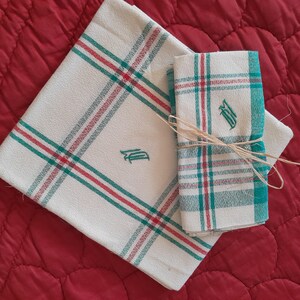 Könnte beinhalten: Zwei weiße Geschirrtücher mit roten und grünen Karomustern. Die Handtücher sind mit dem Buchstaben "D" in grünem Garn bestickt. Die Handtücher sind mit einem Stück Schnur zusammengebunden.