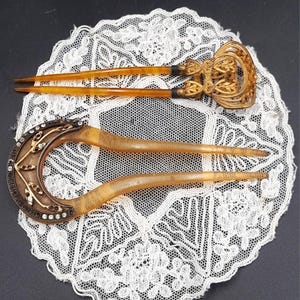 Könnte beinhalten: Zwei antike Haarnadeln. Eine Nadel hat eine dekorative goldfarbene Oberseite mit einem verzierten Design. Die andere Nadel hat eine halbmondförmige Oberseite mit klaren und goldfarbenen Akzenten. Beide Nadeln haben lange, bernsteinfarbene Zinken.