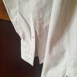 K&ouml;nnte beinhalten: Nahaufnahme eines wei&szlig;en Hemdes mit Kn&ouml;pfen. Das Hemd hat eine gekn&ouml;pfte Manschette und einen Einzelknopfverschluss. Der Stoff scheint mittelschweres Baumwoll- oder Leinenmaterial zu sein, mit sichtbaren N&auml;hten und Steppdetails. Das Hemd ist sauber und knackig.