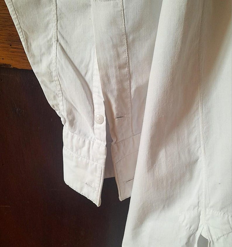 K&ouml;nnte beinhalten: Nahaufnahme eines wei&szlig;en Hemdes mit Kn&ouml;pfen. Das Hemd hat eine gekn&ouml;pfte Manschette und einen Einzelknopfverschluss. Der Stoff scheint mittelschweres Baumwoll- oder Leinenmaterial zu sein, mit sichtbaren N&auml;hten und Steppdetails. Das Hemd ist sauber und knackig.