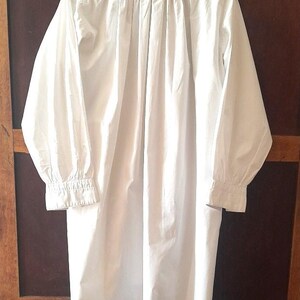 K&ouml;nnte beinhalten: Ein langes wei&szlig;es Hemd mit langen &Auml;rmeln und einem Stehkragen. Der R&uuml;cken des Hemdes hat Falten, die in der Mitte verlaufen. Die &Auml;rmel haben gekn&ouml;pfte Manschetten. Das Hemd ist aus leichtem Stoff.