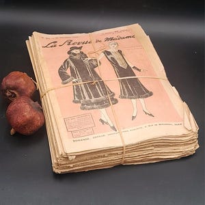 May include: A stack of vintage French fashion magazines, bound with twine. The cover of the magazine, "La Revue de Madame," features illustrations of women in stylish coats and dresses. The pages have a faded pink hue.