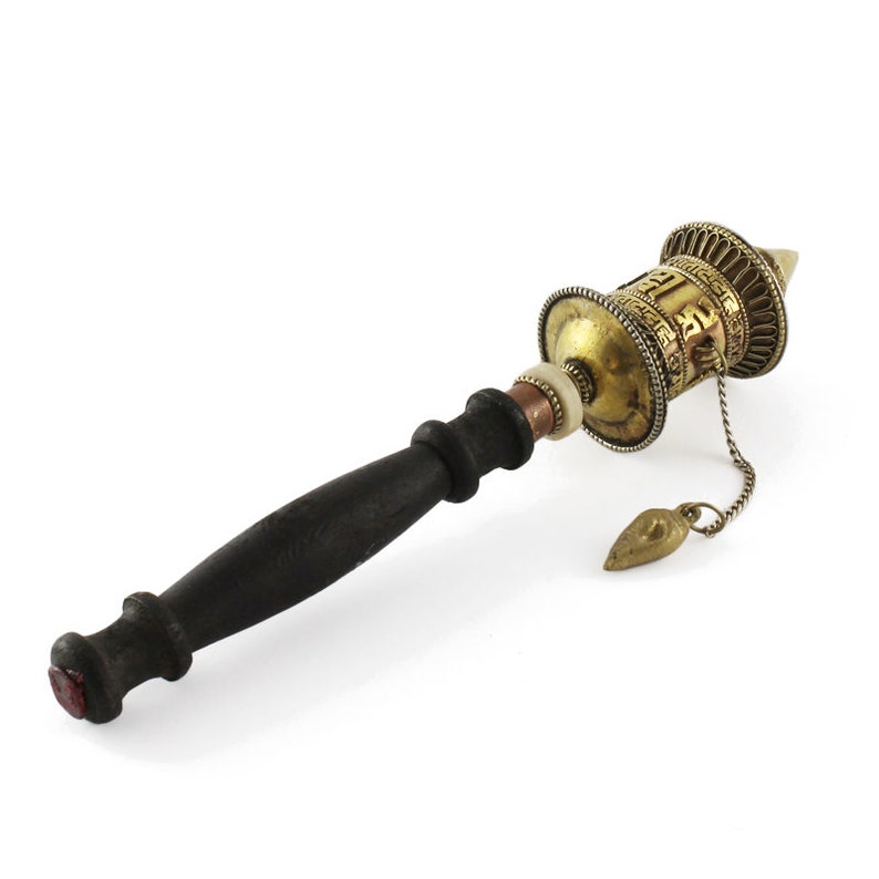 May include: A prayer wheel with a dark wooden handle and a brass cylinder. The cylinder has intricate carvings and is connected to a small brass pendant by a chain. The prayer wheel is a traditional Tibetan Buddhist item.