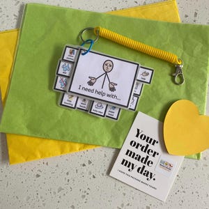 May include: A communication keychain featuring a white card with a stick figure illustration and the text I need help with, attached to a yellow coiled wristband. The set includes smaller cards with icons and a card that says Your order made my day.
