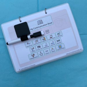 May include: A white communication book with a black dry-erase board and a marker. The book has a spiral binding and features icons for daily routines, including "Lunch Time," "Dinner Time," and "Weekend." The title "My Communication Book" is printed on the cover.