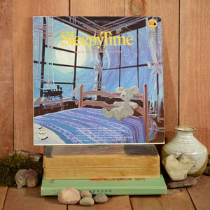May include: A vintage vinyl record album with a blue and white illustration of a teddy bear in bed. The album cover features the title "Sleepytime" and is surrounded by a wooden frame.